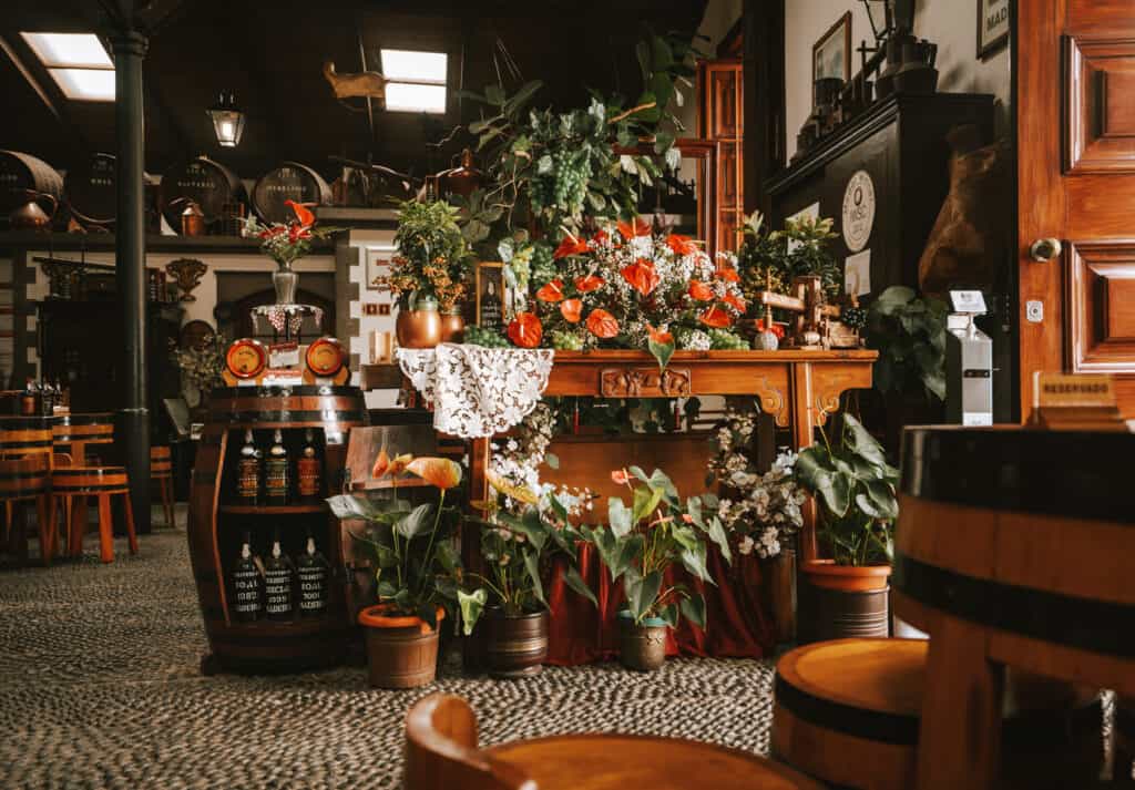 The inside of Pereira D'Oliveira, a famous Madeira winery in Funchal. There are barrels of wine all along the wall and decorative plants and flowers on the tables