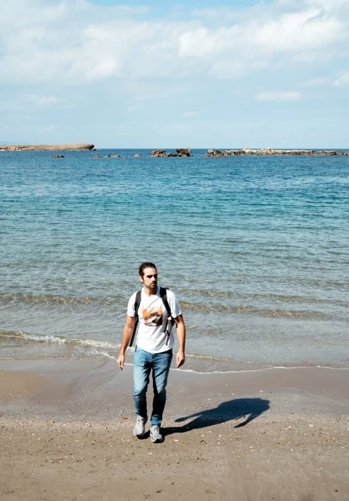 A man with a backpack strolls along the shoreline at Nea Chora Beach, Chania, with calm blue waters and a rocky breakwater in the background, under a clear sky.