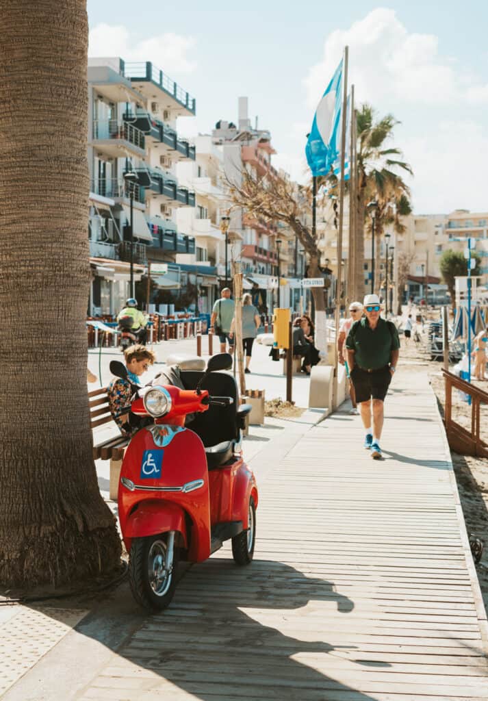 A lively boardwalk at Nea Chora Beach, Chania, with pedestrians strolling by, a red scooter parked on the boardwalk, and the sand leading down the beach