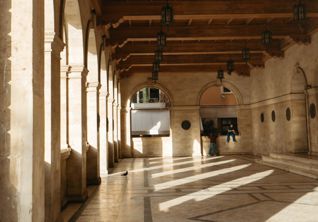 A sunlit colonnade in Heraklion, Crete, featuring tall stone arches and ornate hanging lanterns. The light streaming through the arches creates dramatic shadows on the polished stone floor.