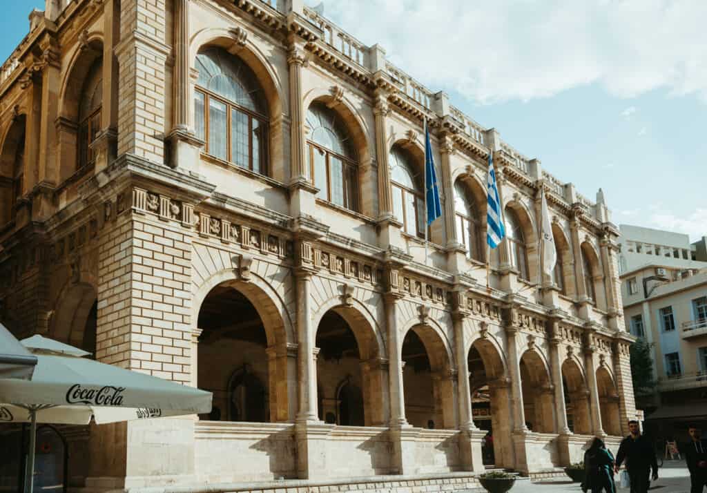 A historic building in Heraklion, Crete, showcasing grand arches and large windows with arched frames on both floors. The facade is adorned with Greek flags waving in the breeze.