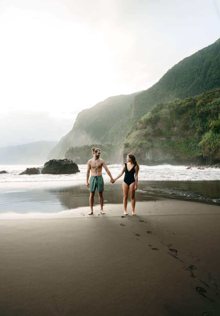 Caleb and Sarah holding hands on Seixal Beach near the ocean. There are mountains in the background