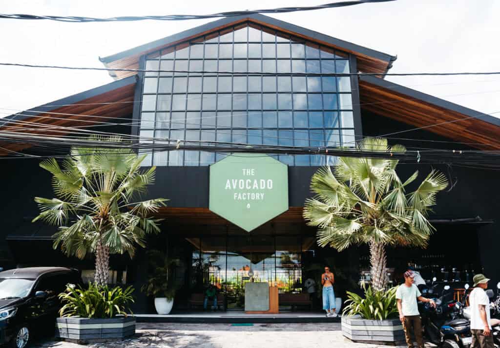 The Avocado Factory brunch in Canggu, Bali, presents a modern façade with a large glass window and a distinctive wooden peaked roof. Its name is displayed on a hexagonal sign against the building's dark exterior, framed by tropical palms, inviting visitors into a space that suggests a focus on fresh and natural cuisine.