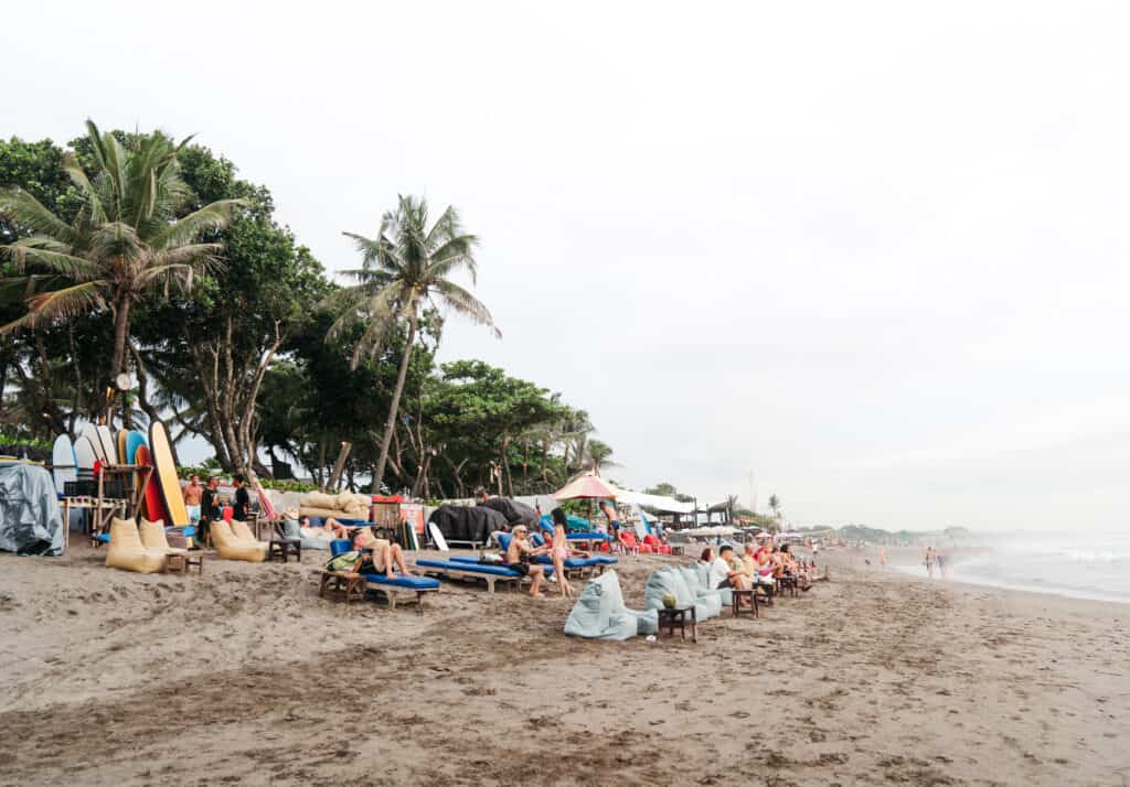 Travelers unwind on beach chairs along the tranquil shores of Canggu Beach, surrounded by lush palm trees and a colorful display of surfboards, embodying the quintessential Balinese beach experience.