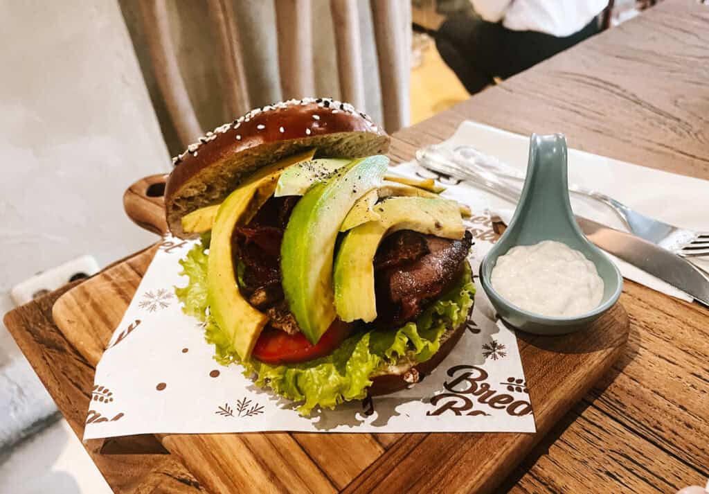 A gourmet burger on a sesame seed bun, presented on a wooden cutting board. The burger is topped with fresh avocado slices, lettuce, and tomato, alongside a spoonful of creamy sauce in a small teal dish. The burger sits on paper with a "Bread Basket" logo, suggesting a casual yet stylish dining experience.