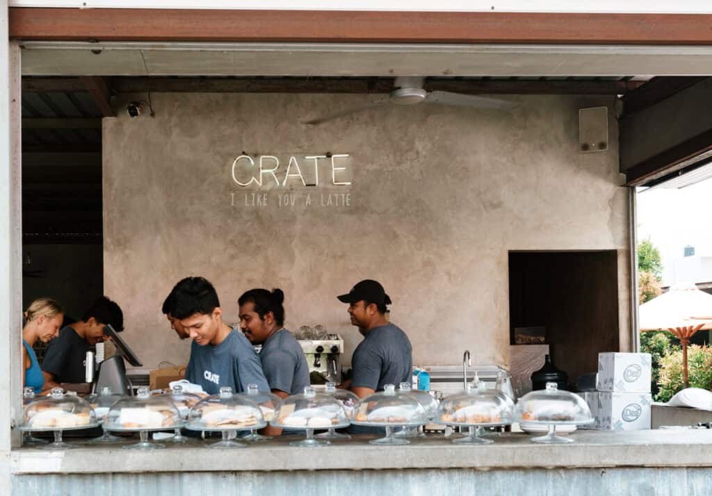 Bustling atmosphere at Crate Cafe with staff wearing branded tees serving customers behind a concrete counter, and an array of fresh pastries under glass domes, under a playful sign that reads 'CRATE – I like you a latte'.