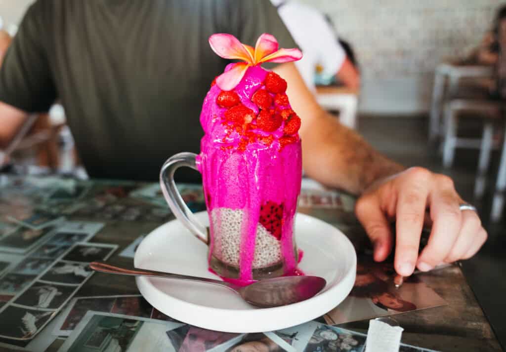A vibrant dragon fruit smoothie topped with a layer of chia seeds and fresh strawberries, adorned with a frangipani flower, served in a clear glass mug on a table covered with a collage of photographs.
