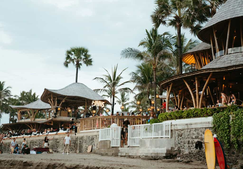 Beachgoers relax at Finns Beach Club in Canggu, Bali, where a neon sign illuminates the unique bamboo structure of the club, with palm trees swaying above and a surfboard propped up in the foreground.