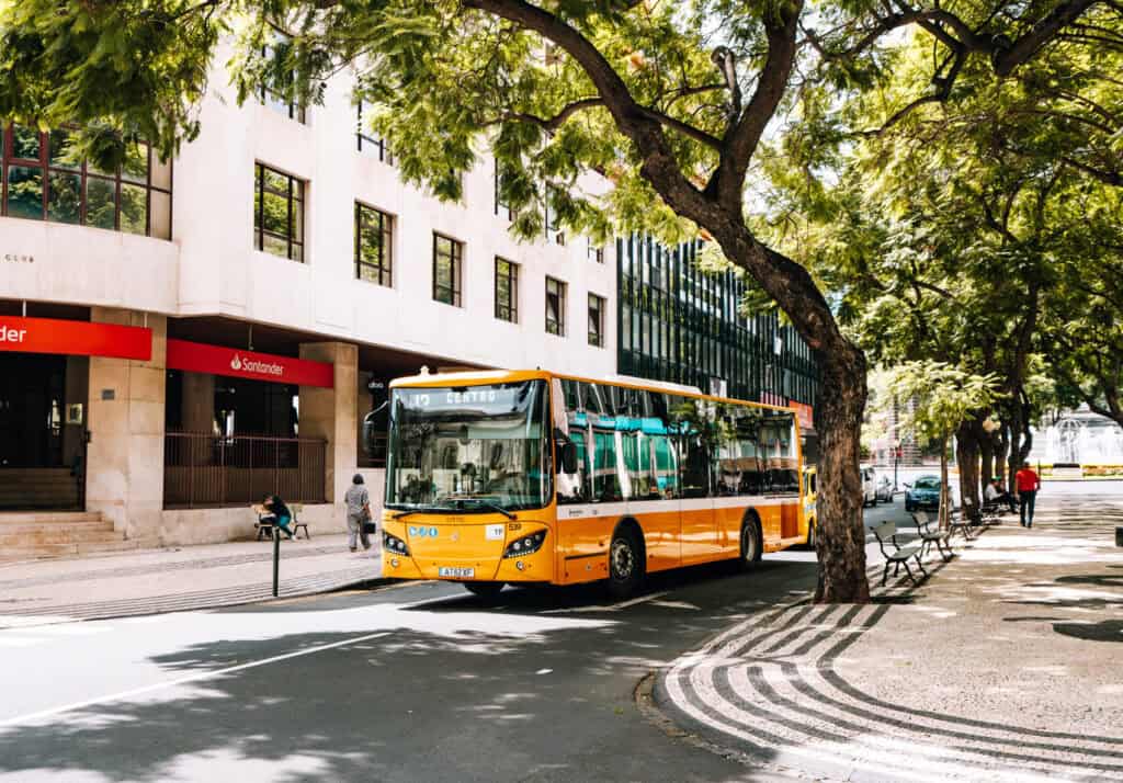 A bright orange public bus labeled 'Centro' stops along a tree-lined avenue in Madeira, with the modern architecture of the city in the background. This street scene, bathed in sunlight, contrasts the public transportation with the convenience of options like Uber in Madeira for getting around the city.
