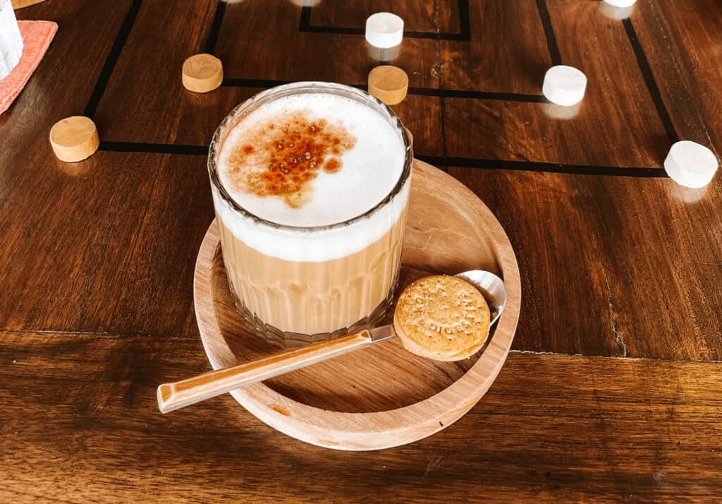 A satisfying cappuccino in a clear glass cup, showcasing the distinct layers of coffee and frothed milk, presented on a round wooden tray. The top is sprinkled with a dash of cinnamon from one of the best places for brunch in Canggu, My Happy Place