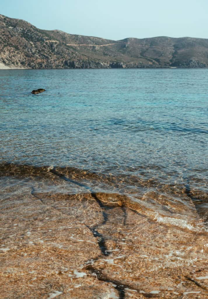 Gentle ripples in the clear waters at the edge of Kedrodasos Beach lead the eye towards a single rock emerging from the sea, with the soft contours of the Cretan mountains stretching across the horizon.