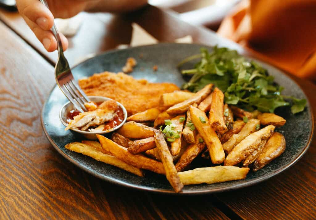 A golden-brown breaded chicken schnitzel, hand-cut fries with a sprinkling of fresh herbs, and a side of mixed greens. The dish is complemented by a small bowl of ketchup, and someone is digging in with a fork, capturing the casual, satisfying experience of a comfort meal.