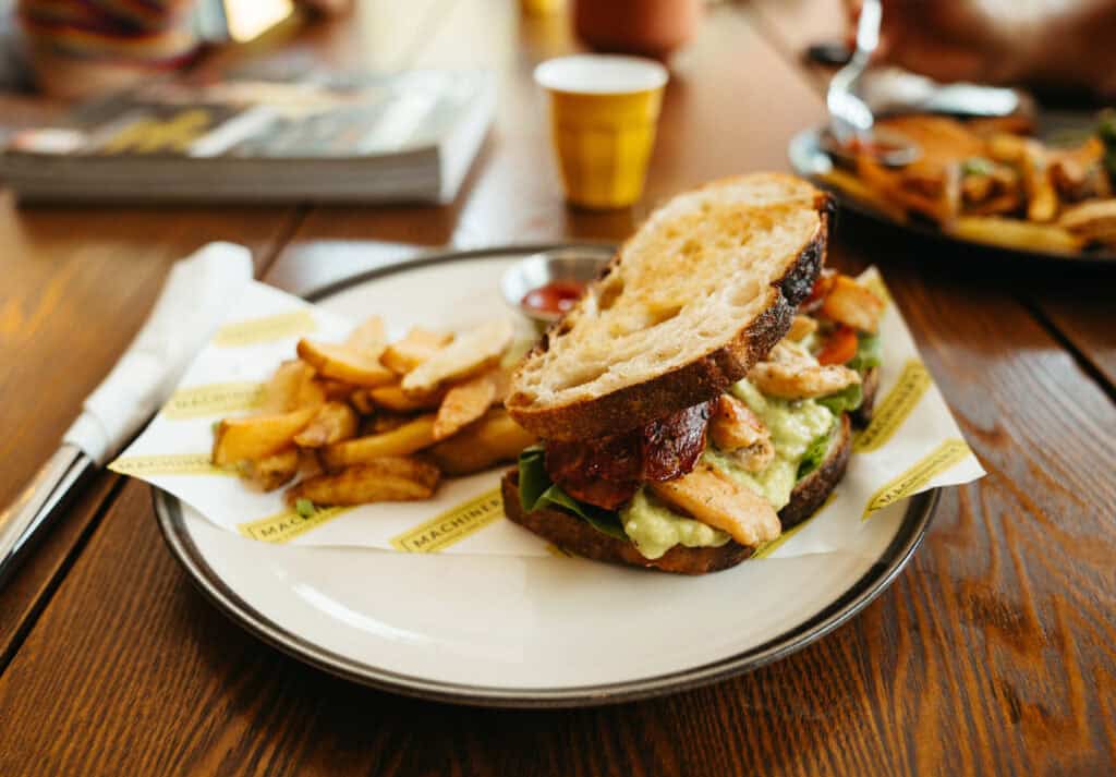 A hearty chicken sandwich on sourdough bread, with a generous spread of avocado, leafy greens, and grilled chicken topped with bacon, served on a round plate. A side of golden, thick-cut fries is ready to be dipped in a small pot of ketchup. The casual dining setting is highlighted by a wooden table and a warm, ambient atmosphere, inviting a relaxed meal for brunch in Canggu.
