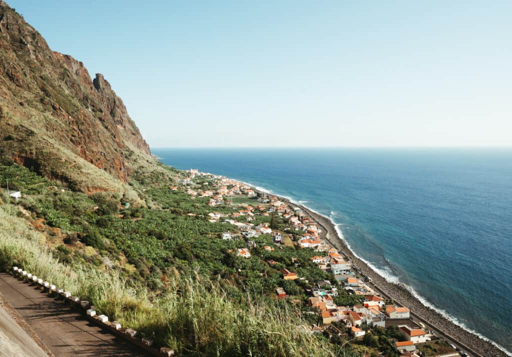 A panoramic view of a coastal village in Madeira, with terracotta-roofed houses nestled among green slopes beside the deep blue sea, as seen from a winding mountain road.
