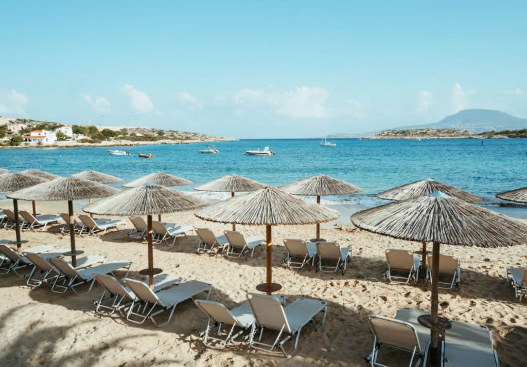 Sun-drenched Marathi Beach with a row of natural straw umbrellas and loungers facing the clear Aegean Sea.