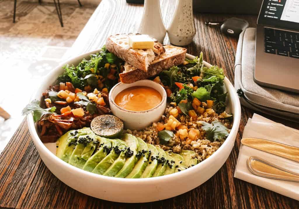 A nourishing bowl of mixed greens, sliced avocado, grilled lemon, corn salsa, toasted bread with butter, and a side of orange dressing, presented on a wooden table beside a laptop, symbolizing a healthy work lunch at Mr. Sista Canggu.