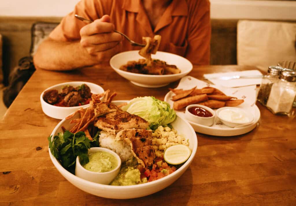 Caleb eating in the background, with focus on a large, healthy meal in the foreground. The dish includes grilled chicken, rice, assorted vegetables, guacamole, and dipping sauces, indicative of a balanced, nutritious offering typical at Nude restaurant in Canggu, Bali.
