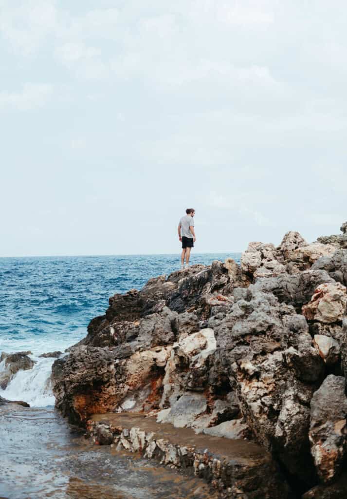 A person in casual summer attire stands atop the natural, rugged rocks of Omprogialos Beach, looking out contemplatively over the vast blue expanse of the Mediterranean Sea beneath a sky dotted with soft clouds.