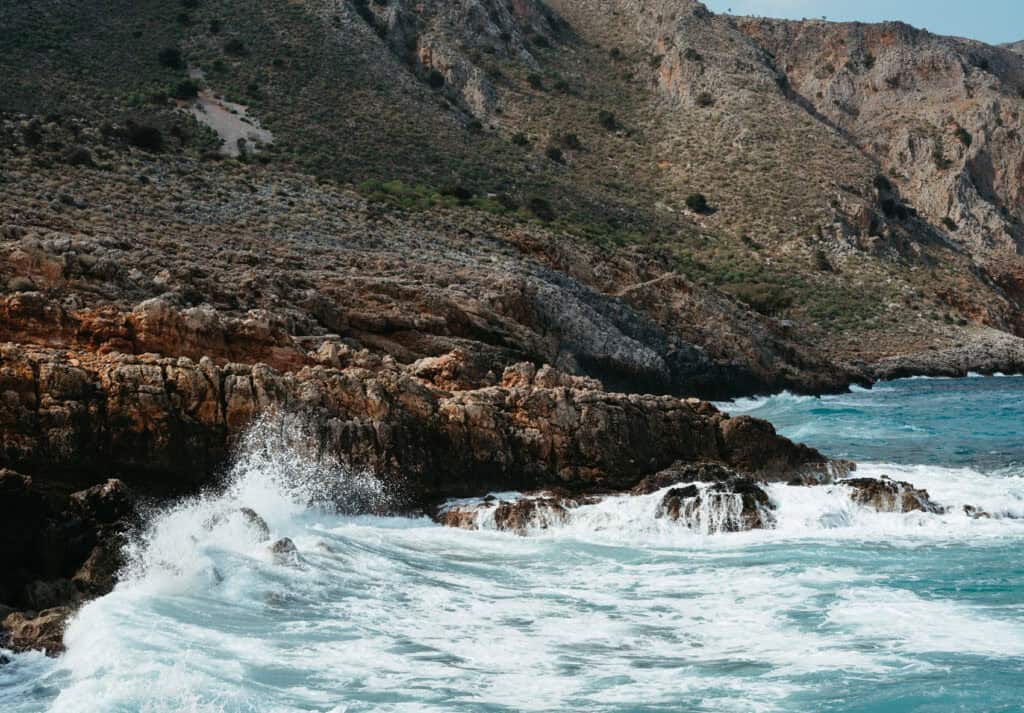 Waves splash energetically against the rocky shores of Omprogialos Beach, with the contrasting calmness of the sea and the ruggedness of the Cretan mountains in the background, highlighting the wild beauty of this coastal retreat.