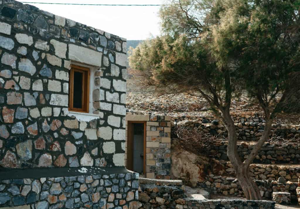 A traditional stone house near Omprogialos Beach with a small wooden window, set against a backdrop of a dry stone wall and a resilient tree, capturing the rustic charm of the Cretan landscape.