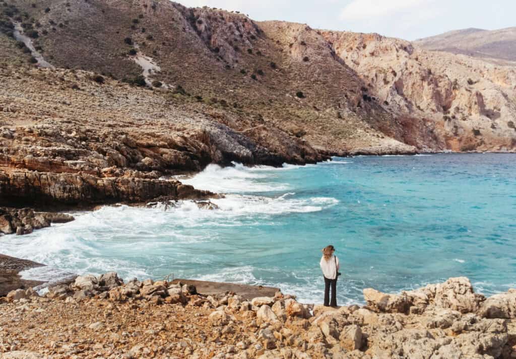 Sarah stands on the pebbly shore of Omprogialos Beach, gazing at the clear turquoise waters that contrast with the rugged mountains of Crete.