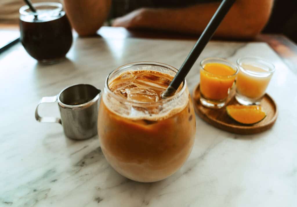A cold brew coffee with milk swirls served in a glass jar with a metal straw, accompanied by two small shots of ginger juice on a wooden tray, all set on a white marble tabletop, creating a refreshing scene for a coffee break.