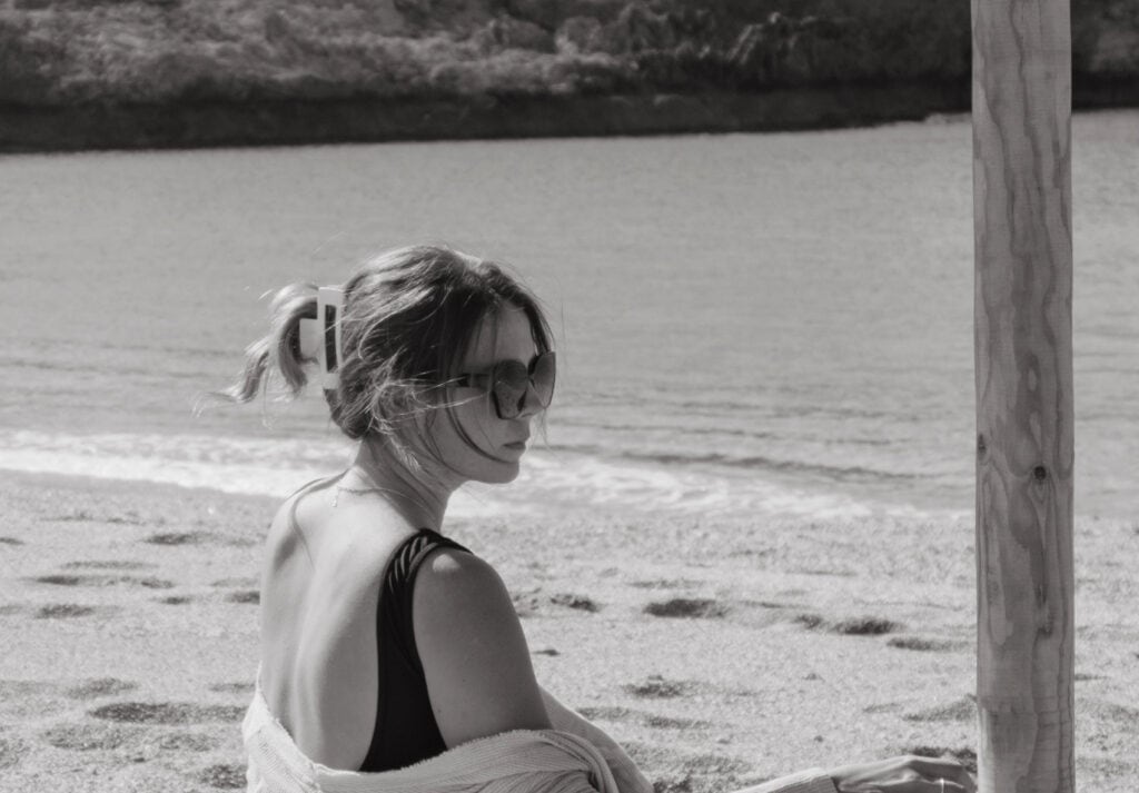 Black and white photo of Sarah sitting on a beach chair at Skinaria Beach, Crete. She is partially wrapped in a towel, wearing sunglasses, and looking away from the camera towards the sea, with a wooden pole partially framing the shot. The sandy beach and calm sea create a serene backdrop.