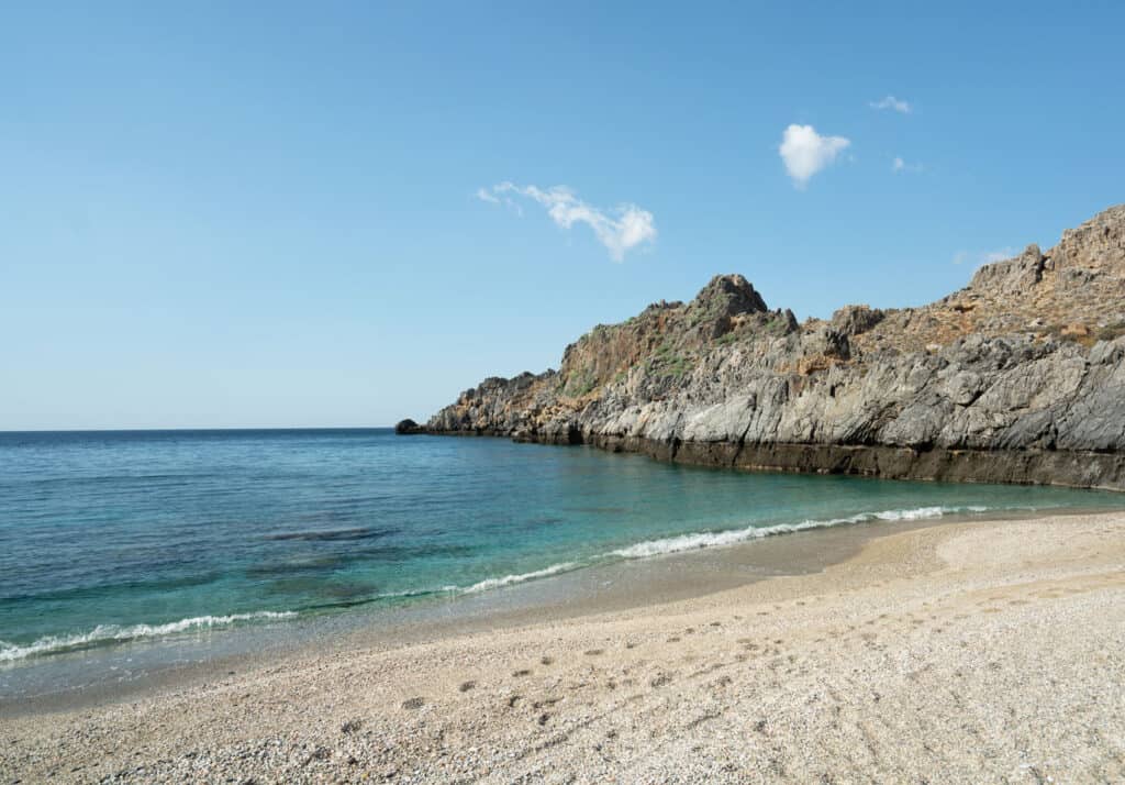 The pristine waters of Skinaria Beach lap gently at a pebbly shore, bordered by dramatic rocky cliffs under a clear blue sky, showcasing the natural beauty and tranquility of this secluded Cretan beach.