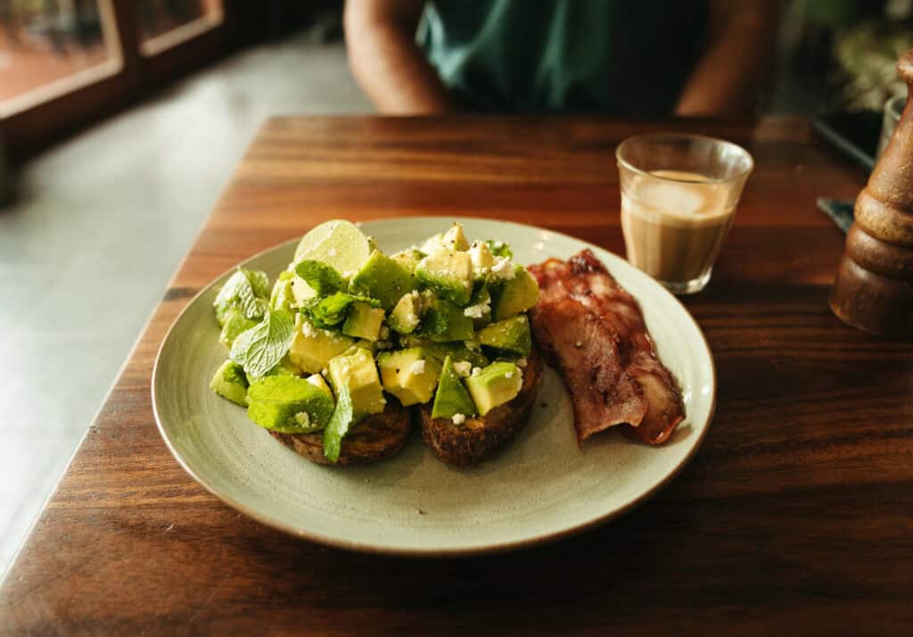 A close-up view of a hearty breakfast in Canggu, featuring avocado toast topped with chunks of avocado, crumbled cheese, and a sprinkle of herbs. Accompanying the toast are two crispy bacon strips and a small glass of espresso. The warm, ambient light highlights the texture of the food and the natural wood grain of the table.