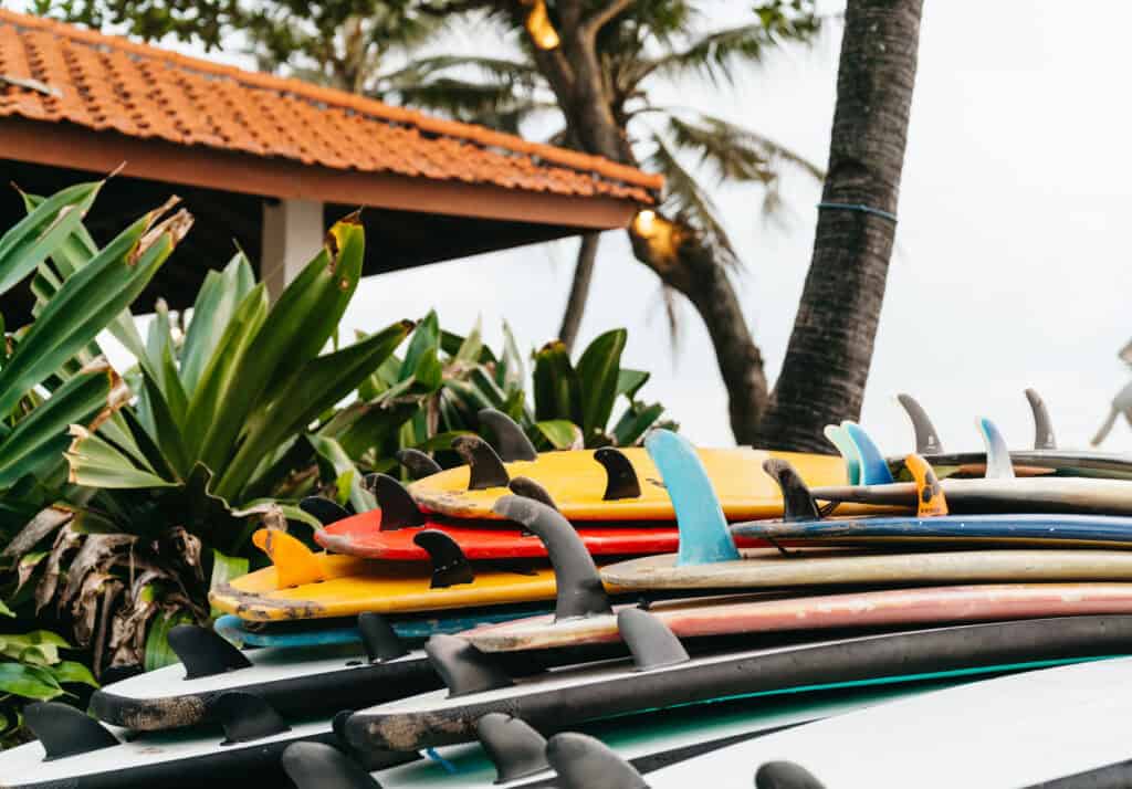 A stack of colorful surfboards awaits eager surfers against a backdrop of tropical foliage and a traditional Balinese rooftop, capturing the surf culture of Canggu Beach.