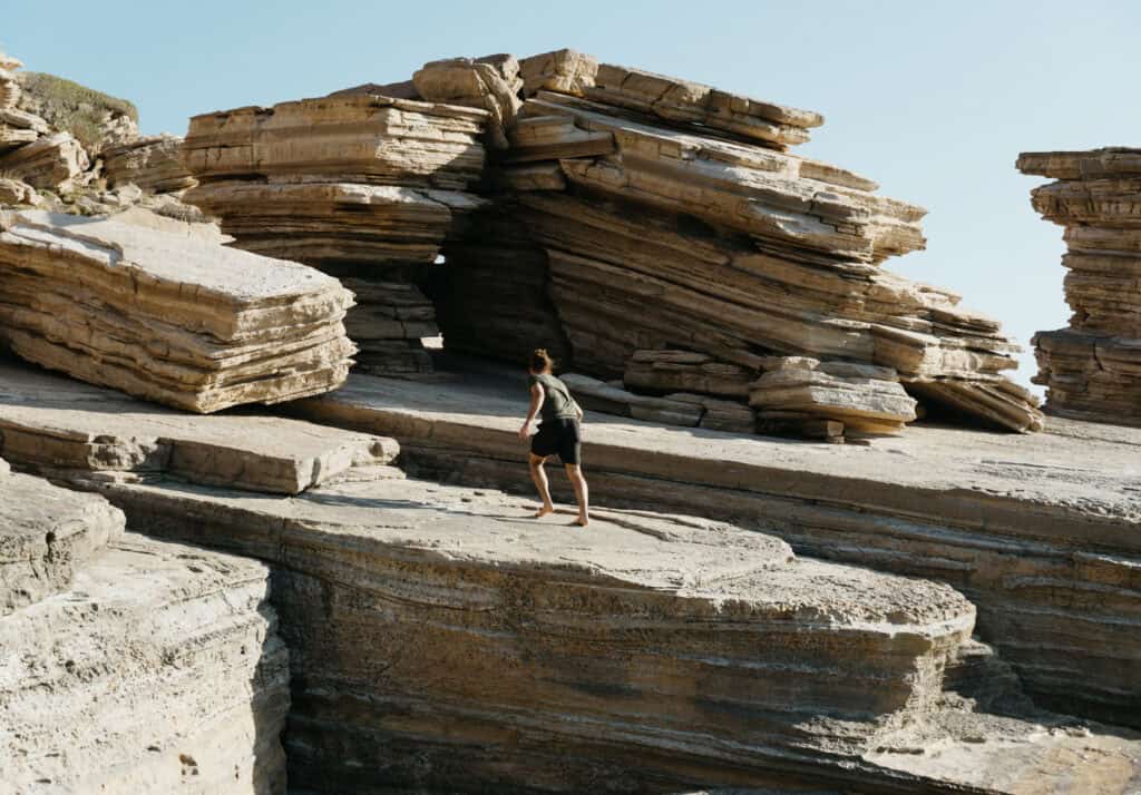 Caleb explores the layered rock formations at Triopetra Beach on Crete. The geological structures feature multiple stacked layers of sedimentary rock, creating rugged caves and archways. The sun casts sharp shadows on the textured stone surfaces, emphasizing the natural beauty of the coastal landscape.