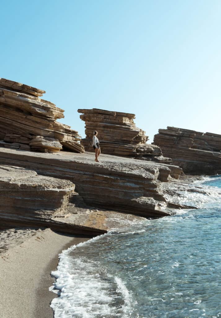 Sarah walks along the layered rock formations at Triopetra Beach, Crete. The rock layers appear in horizontal strata, creating dramatic natural shelves leading down to the clear blue waters. The sunlit scene captures the tranquil yet rugged beauty of the beach, highlighted by the pristine sandy shore and gentle waves lapping at the rocks.