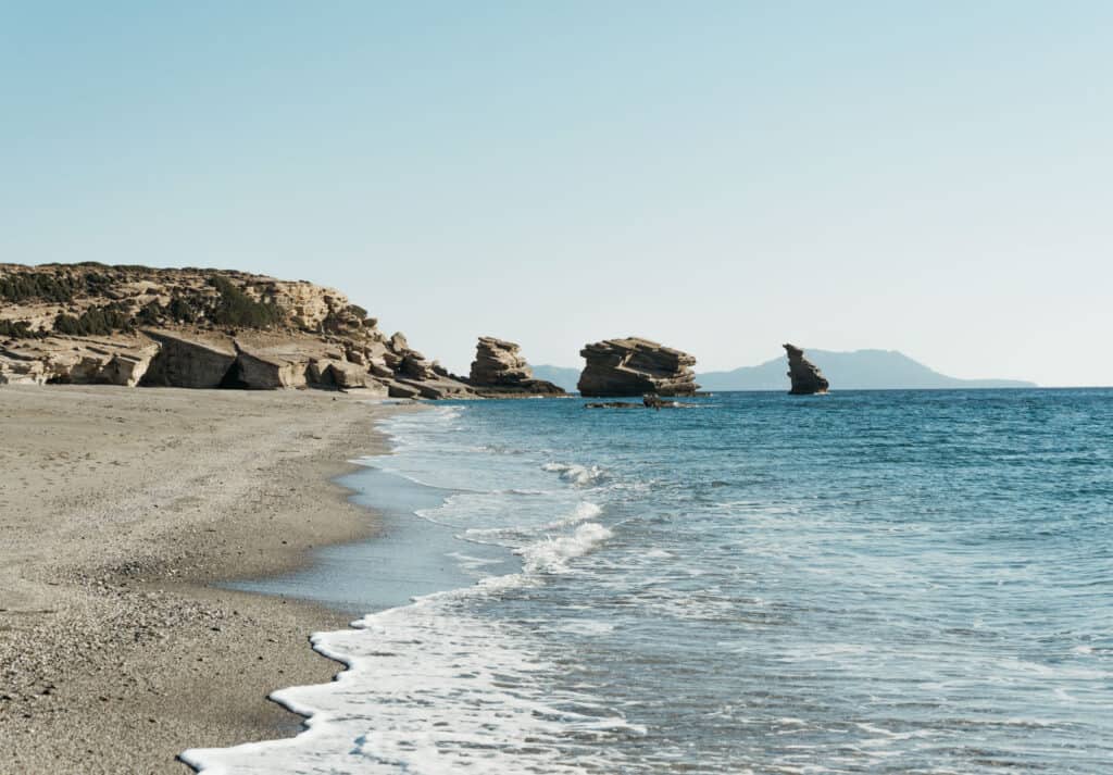 A serene view of Triopetra Beach featuring the beach's distinctive layered rock formations along the shoreline. The clear blue waters gently lap at the sandy beach, with the silhouettes of distant mountains under a bright blue sky. The scene is quiet and untouched, capturing the natural beauty of this remote location.