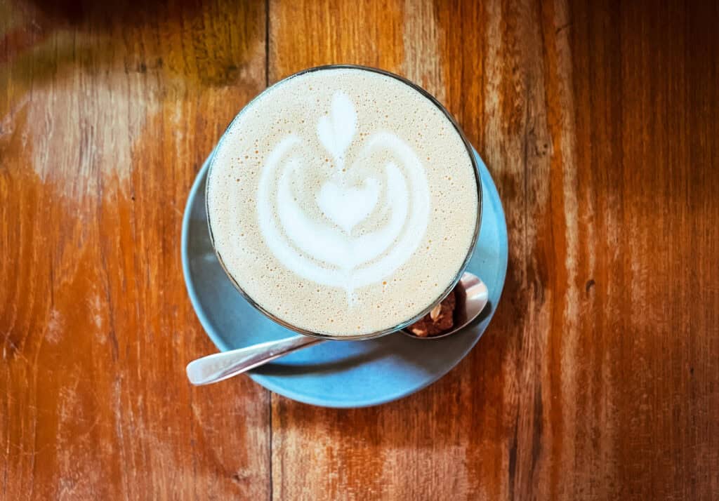 Top-down view of a freshly made cappuccino with artistic latte art on a wooden table. The creamy foam boasts beautiful latte art, accompanied by a crisp cookie on the saucer and a spoon to the side, capturing a cozy café ambiance.