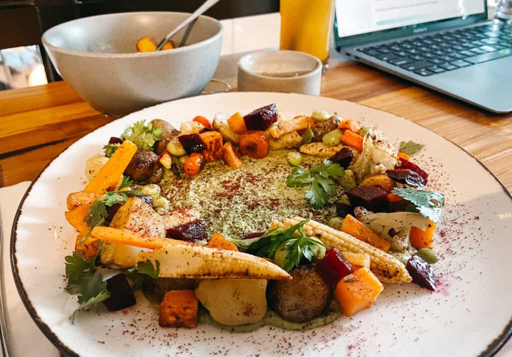 A vibrant plate of roasted vegetables, including beets, carrots, and corn, garnished with fresh herbs, served on a speckled plate. In the background, a grey bowl, a glass of juice, and a laptop open on a wooden table hint at a productive brunch in Canggu at Zin Cafe.