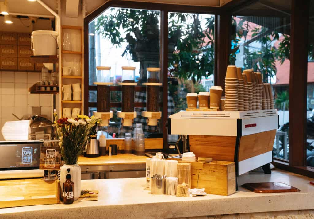The counter of Baked café in Canggu, adorned with fresh flowers and filled with coffee essentials, alongside a pristine coffee machine and stacked takeaway cups, offering a glimpse into the vibrant café culture.