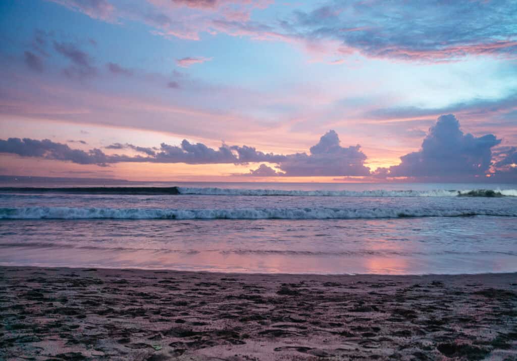 A serene sunset at Canggu Beach with pastel pink and blue skies reflected on the water's surface. Waves gently approach the sandy shore, creating a picturesque and peaceful evening scene.