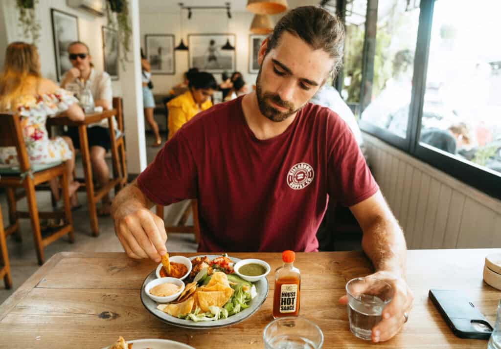 Caleb enjoys a colorful plate of food with dips and a side of hot sauce at Commons Canggu, a bustling café with other patrons in the background, encapsulating a casual dining experience.