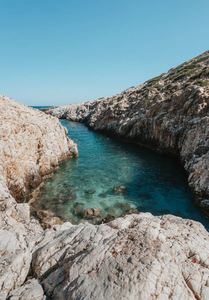 Crystal-clear turquoise waters flow through a narrow channel flanked by textured limestone cliffs under a bright blue sky, capturing the serene beauty of Katholiko Beach, Crete.