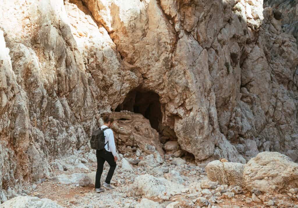 Caleb with a backpack explores the rugged terrain near the entrance of a cave in the limestone cliffs of Katholiko, Crete, bathed in the warm light of the sun.