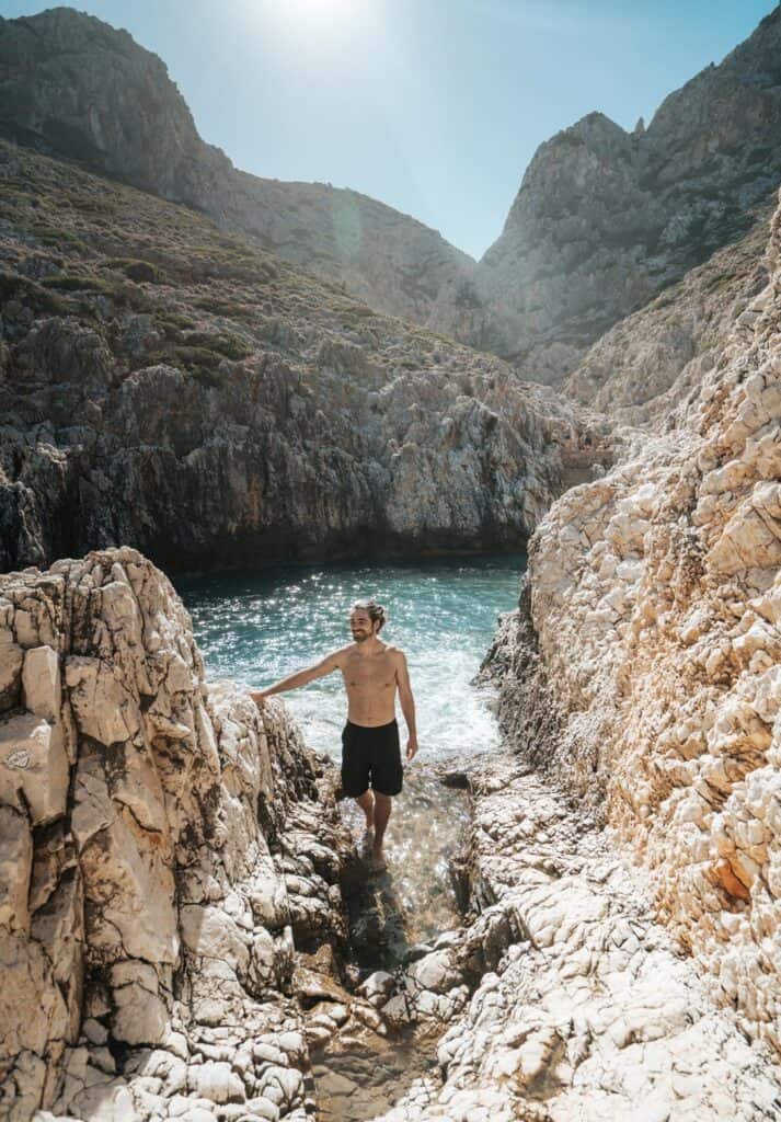 Caleb in black shorts smiles as he stands on a craggy limestone shore, with the clear blue waters of Katholiko beach behind him