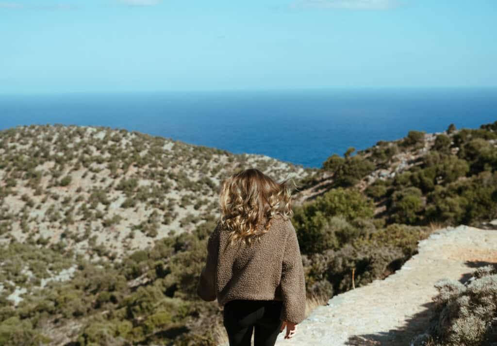 Sarah gazes out over a serene landscape of rolling hills and Mediterranean vegetation leading to the vast blue sea under a clear sky.