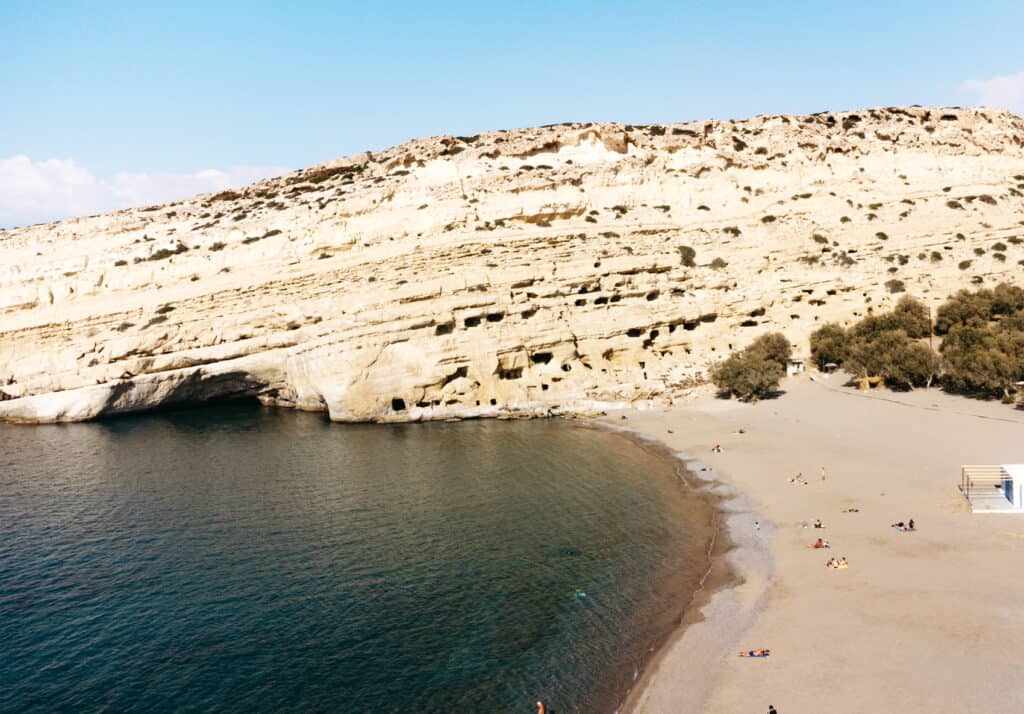 Aerial view of Matala beach in Crete, showcasing the golden sandy shore lining the clear blue waters of the Mediterranean Sea, with a backdrop of weathered cliffs peppered with ancient cave openings.
