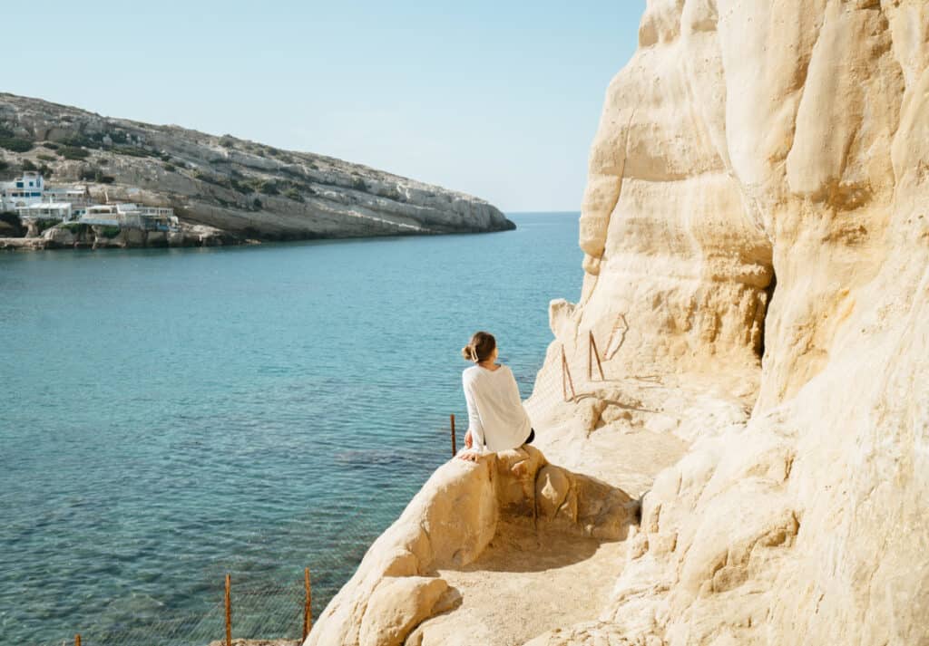 Sarah sits on a rocky outcrop in Matala, Crete, looking out over the serene blue sea, with the sandy cliffside curving into the distance. The scene is bathed in bright sunlight, highlighting the rugged texture of the cliffs and the peaceful expanse of water.