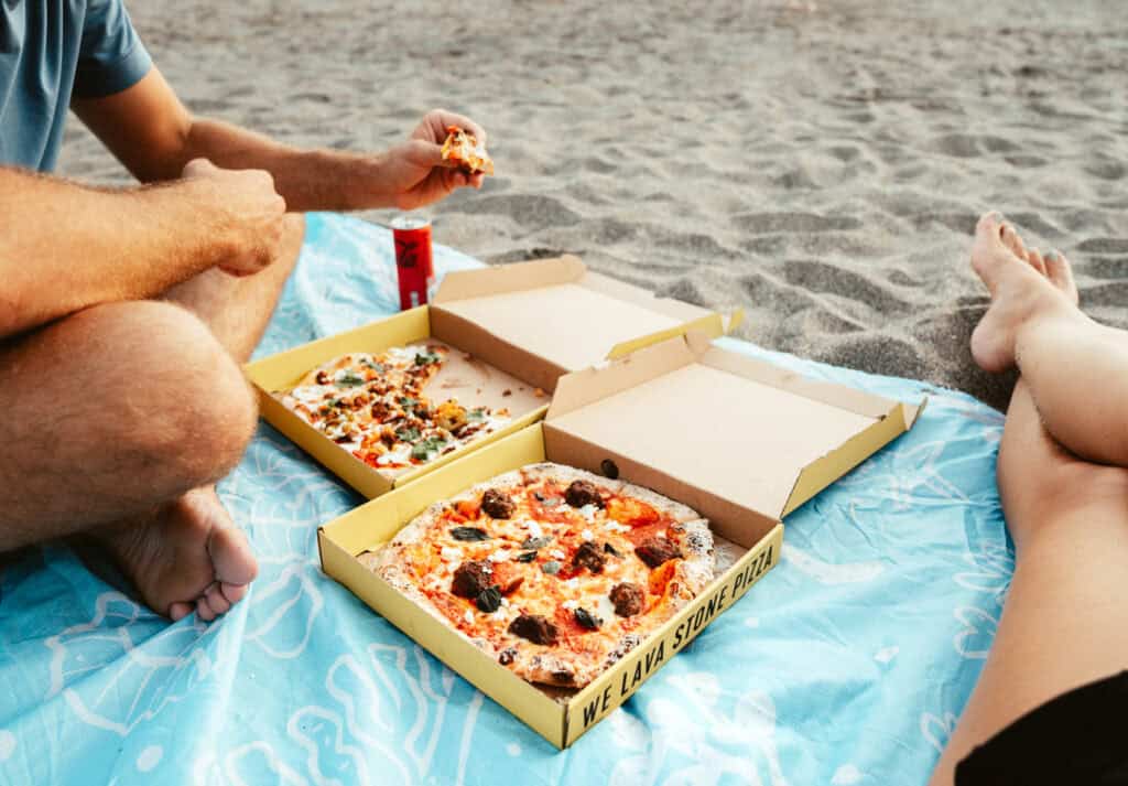 A beach pizza picnic is among the relaxing things to do in Canggu, featuring Caleb and Sarah enjoying freshly made pizzas on a blue picnic blanket, with a view of the sandy beach and ocean at their feet.