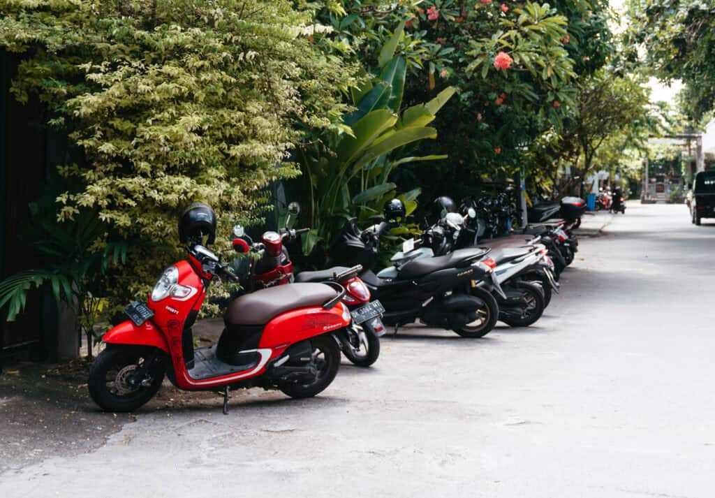 A row of parked scooters, led by a vibrant red one with a helmet on its handle, lines the side of a serene street in Canggu, framed by lush tropical foliage. Renting a scooter in Bali is an excellent way to get around.