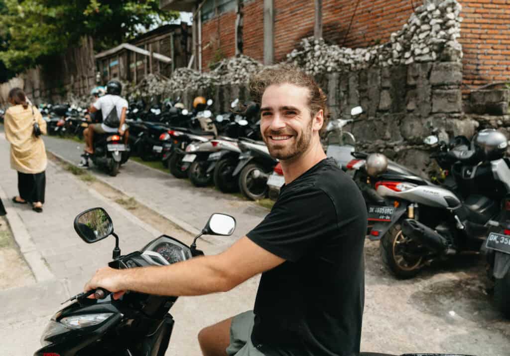 Caleb sitting on a black scooter, ready to ride on a street lined with parked motorbikes in Canggu. The background features a brick wall with climbing plants, adding to the laid-back, urban atmosphere.