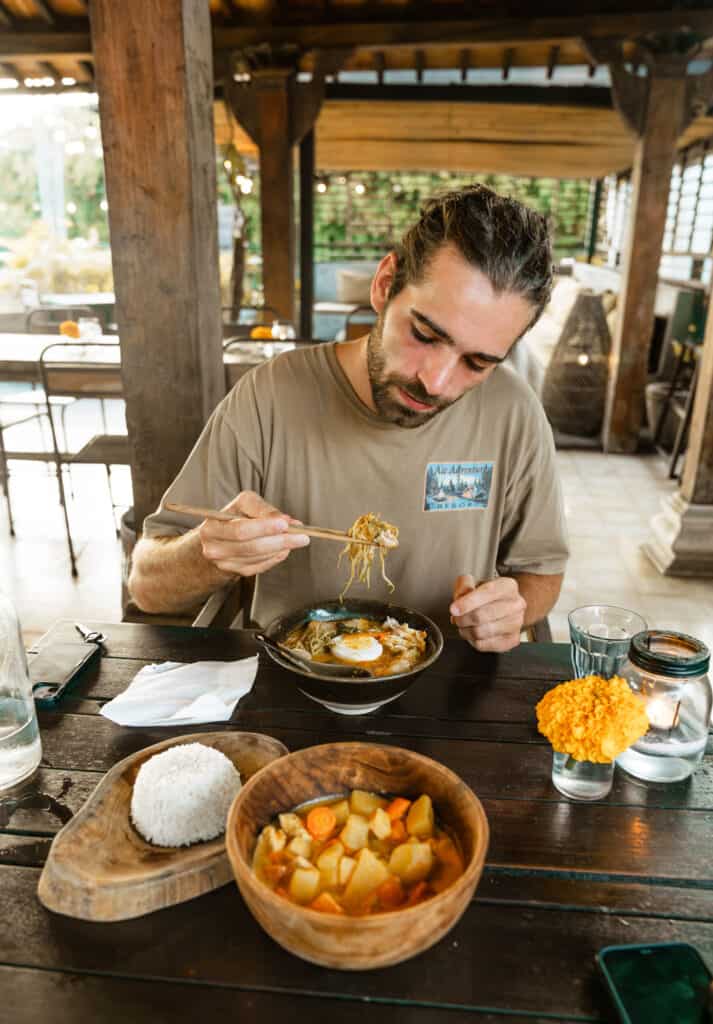 Caleb is enjoying a meal at a wooden table in a rustic, open-air dining setting. He's eating a bowl of noodle soup topped with a fried egg, with a side of white rice shaped into a dome on a wooden plate and another bowl of vegetable curry, creating a relaxed and appetizing dining experience.