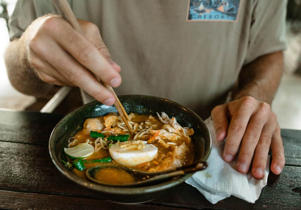 Caleb handles chopsticks to eat from a bowl of rich, aromatic ramen with a half-boiled egg, fresh greens, and bean sprouts from Warung Ithaka, one of the best warungs in Canggu, Bali