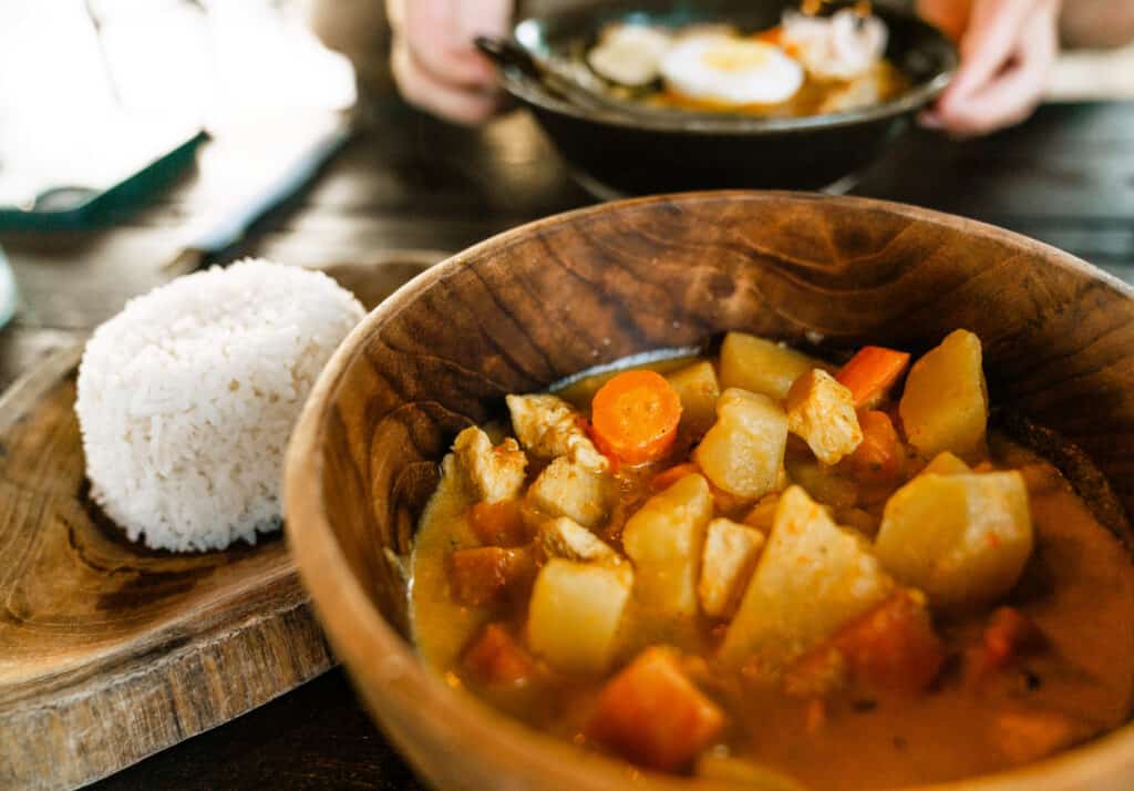 A close-up view of a traditional Indonesian dish, with a neatly shaped dome of white rice beside a wooden bowl of hearty chicken curry, featuring tender meat, potatoes, and carrots.